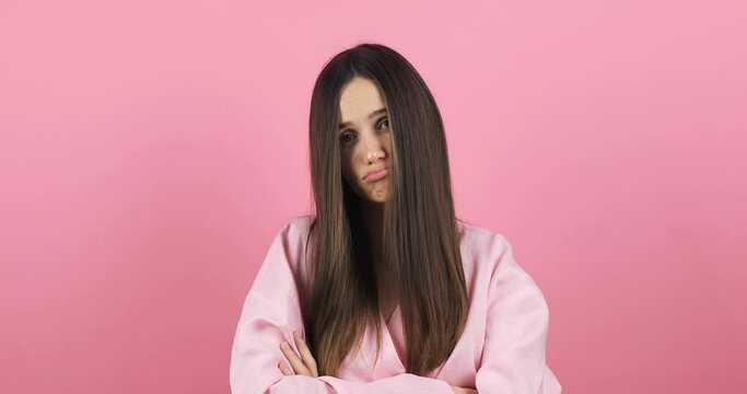 Upset brunette hair woman wear pink dress purses lips offendedly and looks from under forehead on pink background. Sad tired woman looks with poker face at camera purses lip.