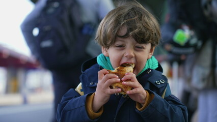 Portrait of a delighted happy child eating croissant standing at train platform wearing jacket and...