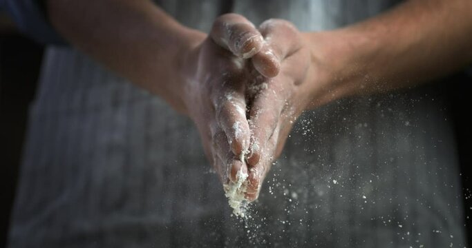 Super slow motion macro of professional artisan baker claps his hands with flour while prepare a dough for making homemade bread or pizza on rustic wooden table in traditional bakery.