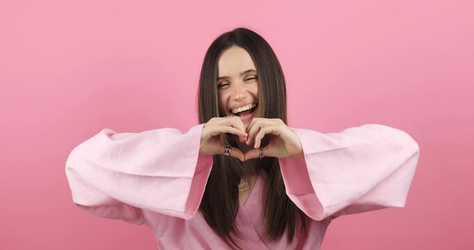 Happy attractive brunette woman look camera showing shape heart with hands, girl wear pink dress isolated on pink background. Love, romantic concept.