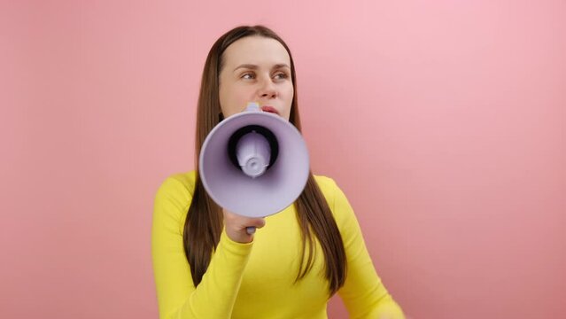 Angry young woman loudly screaming holding speaker in hand, strict boss yelling, raising fist, wearing yellow sweater, posing isolated on pink color background wall in studio. Solving problem concept