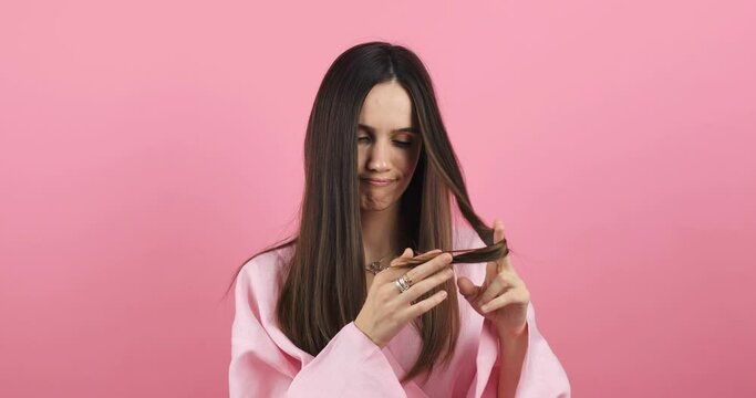 Depressed young woman looking at split brunette hair ends. Worried sad girl feels upset about brittle damaged dry hair loss concept. Female hormone problems concept isolated pink background.
