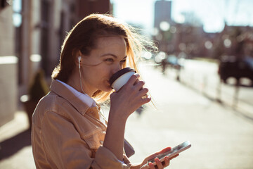 Smiling woman walking and drinking coffee, happy female with smartphone, urban lifestyle, casual...