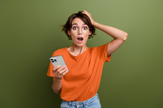 Photo of speechless crazy girl dressed orange t-shirt hold smartphone arm on head astonished staring isolated on khaki color background