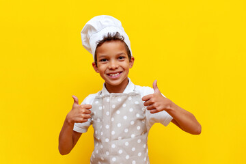 african american boy in chef's uniform and hat shows likes on yellow isolated background, teenager...