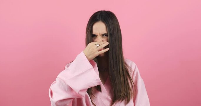 Disgusting odor. Discontent brunette woman frowning face, smells something awful, feeling aversion, pink studio background. Girl face with aversion dislikes something unpleasant. Woman wear pink dress