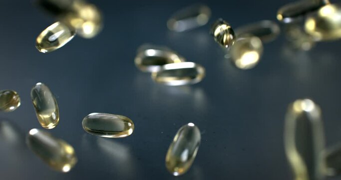 Slow motion macro of transparent yellow soft gel vitamins and nutritional supplements capsules falling on grey mirroring surface in scientific pharmaceutical laboratory.