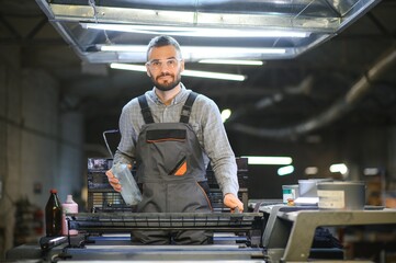 Printing house interior with modern offset print machine and operator in working uniform checking...