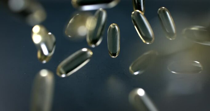 Slow motion macro of transparent yellow soft gel vitamins and nutritional supplements capsules falling on grey mirroring surface in scientific pharmaceutical laboratory.