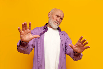 old bald grandfather in purple shirt refuses and shows stop gesture on yellow isolated background
