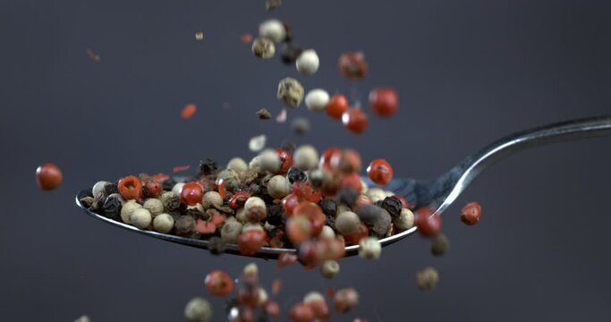 Super slow motion macro of dry mixed flavorful and aromatic peppercorn falling down from silver spoon isolated on soft dark background.