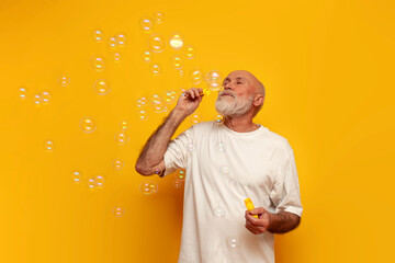 cheerful old bald grandfather in white t-shirt blowing soap bubbles on yellow isolated background