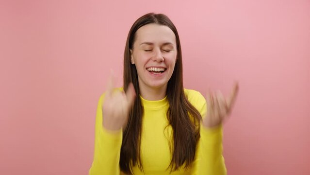 Portrait of overjoyed young woman showing rock and roll hand sign, screaming and gesturing to heavy metal, rock music, wearing yellow sweater, posing isolated over pink color background wall in studio