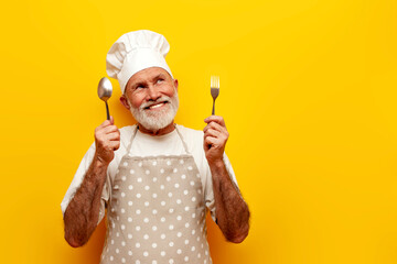 pensive old grandfather chef in hat and apron holds fork and spoon and dreams on yellow isolated...