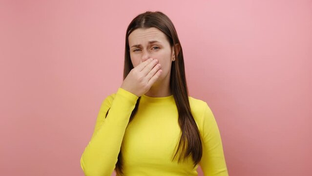 Bad smell concept. Upset young woman standing pinching her nose with fingers to hold breath, disgusted by stinky intolerable smell, wearing sweater, posing isolated on pink background wall in studio