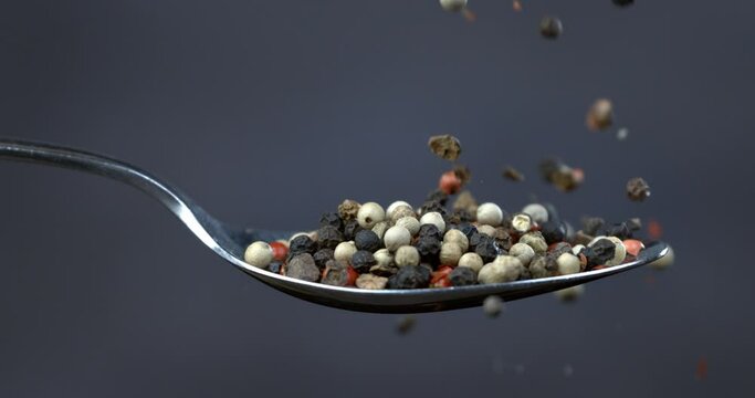Super slow motion macro of dry mixed flavorful and aromatic peppercorn falling down from silver spoon isolated on soft dark background.