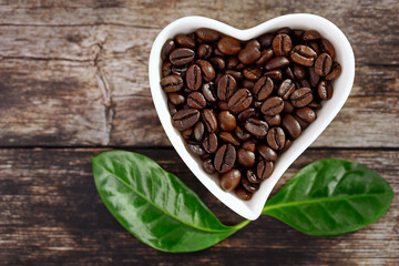 top view of roasted coffee beans in white heart-shaped bowl on wooden background with green leaves of coffea arabica, concept for coffee lovers