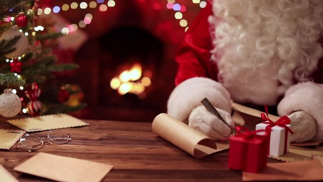 Santa Claus writes while sitting at a table near a fireplace with fire