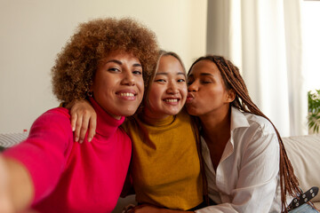 African American women and Asian woman sitting on sofa at home taking selfie and hugging