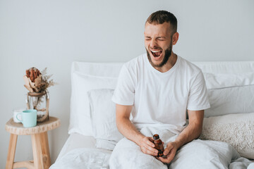 Sleepy young caucasian man in bed yawns holds jar with seeping pills, feels fatigue, needs healthy...