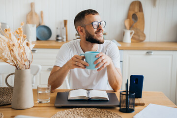 Cheerful caucasian bearded man in glasses sitting at table holds cup of coffee looks away happily, thinking about career, remote works with laptop, planning agenda. Purposeful guy remote working.