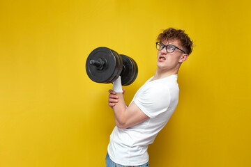 young nerd guy in glasses holds heavy dumbbells and strains on a yellow background, a motivated guy...