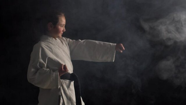 Young karate girl wearing kimono and posing against black background with smoke
