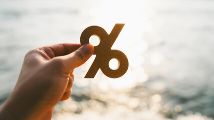 Businessman's hand holding a symbol Percentage with blur sea background, concept of systems of raising or lowering Fed interest rates to correct inflation concepts.