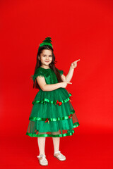 A girl in a festive carnival costume of little Christmas tree pointing at an empty space on a red background. Winter holidays concept. Sale and advertising