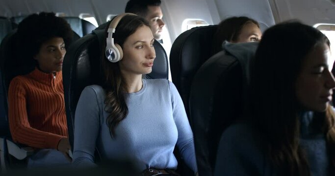 Young woman with headphones listening her favorite music and relaxing with comfort on board of aircraft while flying at airplane to holidays vacation destination.
