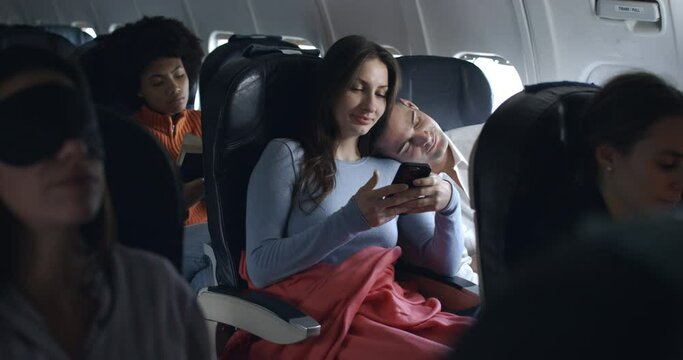 cute couple of friends in airplane have a nice moment together   watching pictures, video call and smiling with sleepy boyfriend during the flight from the distant airport.