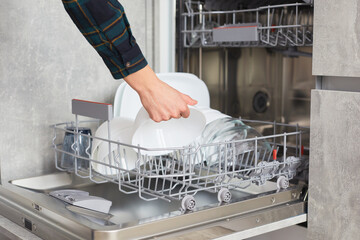 A woman's hands load and and takes out plates, cups and other dishes into an open dishwasher in a modern bright kitchen. The concept of household home life