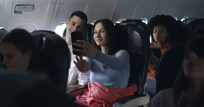 Cute couple of friends in airplane have a nice moment together and having fun taking and watching pictures, video call and laughing  during the flight from the distant airport.