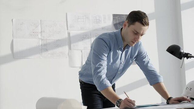 Engineer working in a bright office, standing in front of the table. A young man in a shirt with a wrist watch. Design and work with drawings.