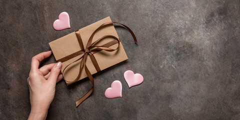 Female hand holds a brown envelope tied with a silk ribbon and pink silk hearts on a dark vintage background. Valentine's Day card. Top view, flat lay, copy space. Banner - obrazy, fototapety, plakaty