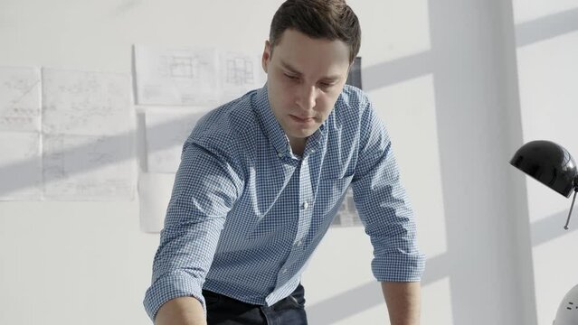 Architect draws a blueprint, standing in front of the table. Light workspace, helmet on the desk. General plan, shooting from side.