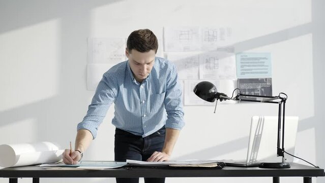 Male designer 30-35 years old working on a project. He stands near the table, makes notes. Bright minimalist studio and drawings on the wall.