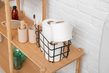 Basket with toilet paper rolls and bath supplies on wooden shelving unit near light brick wall,...