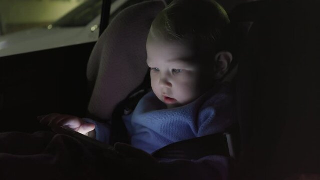 portrait of a small child sitting in a car at night in a child seat and looking at a smartphone. High quality 4k footage
