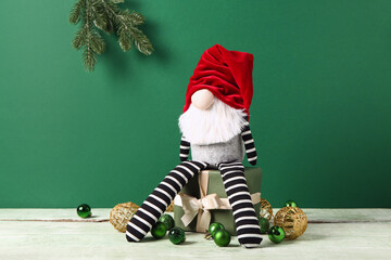 Christmas gnome with gift box, balls and fir branch on wooden table against green background