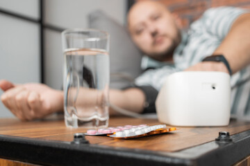 Defocused background with man who has hypertension and headache, lying on sofa at home, measures blood pressure on hand with modern digital tonometer, focus on blister pack with pills on foreground