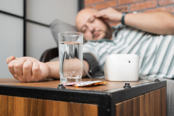 Obraz na płótnie Canvas Defocused background with man who has hypertension and headache, lying on sofa at home, measures blood pressure on hand with modern digital tonometer, focus on blister pack with pills on foreground