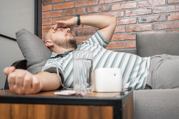 Unhealthy young man suffering from hypertension and headache, lying on the sofa at home, measures...