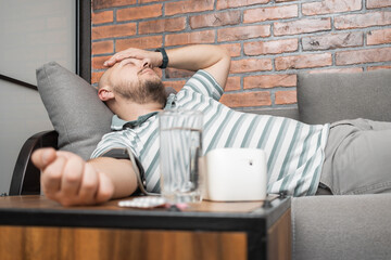 Unhealthy young man suffering from hypertension and headache, lying on the sofa at home, measures...
