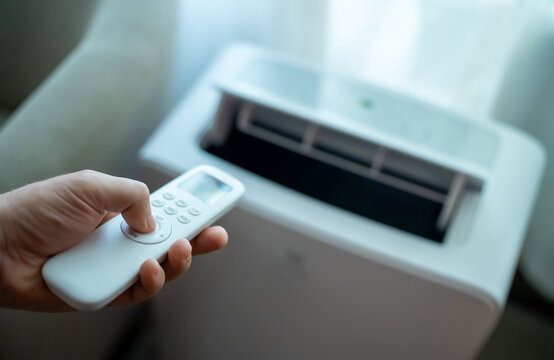Hand holding remote control aimed at floor standing mobile air conditioner.