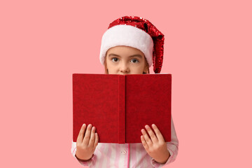 Cute little girl in Santa hat with book on pink background
