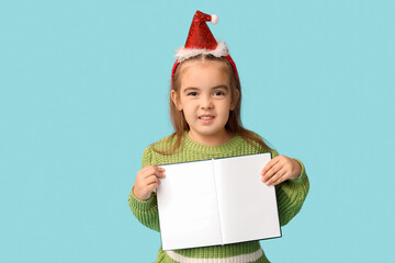 Cute little girl in Santa hat headband with book on blue background