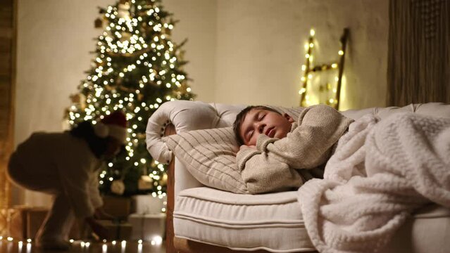 Mother secretly putting Christmas box under illuminated xmas tree while son sleeping on a light sofa in a cozy room with muffled light and glowing garlands. High quality 4k footage