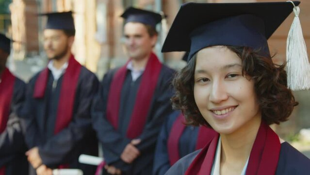 Close up portrait of cheerful female bachelor in hat and gown looking at camera, standing near other students listening to valedictory