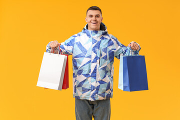 Young man in stylish puffer jacket with shopping bags on yellow background